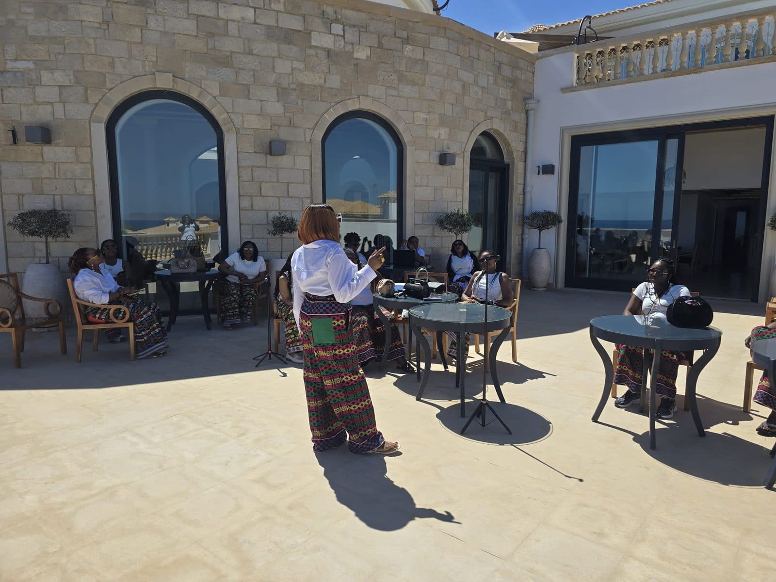 A woman speaking to a seated group of women on a sunlit terrace