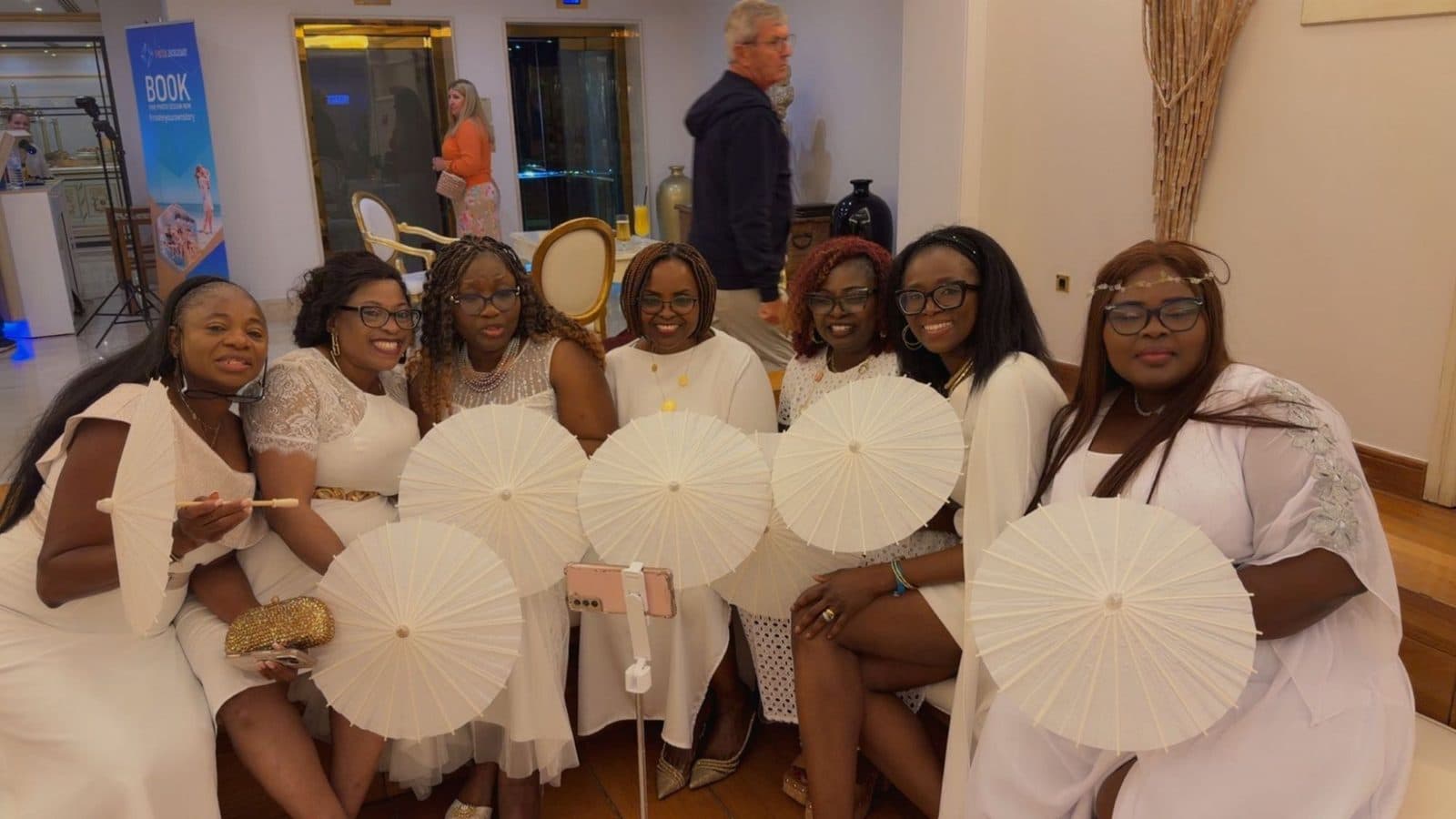 Seven women in white dresses holding paper parasols